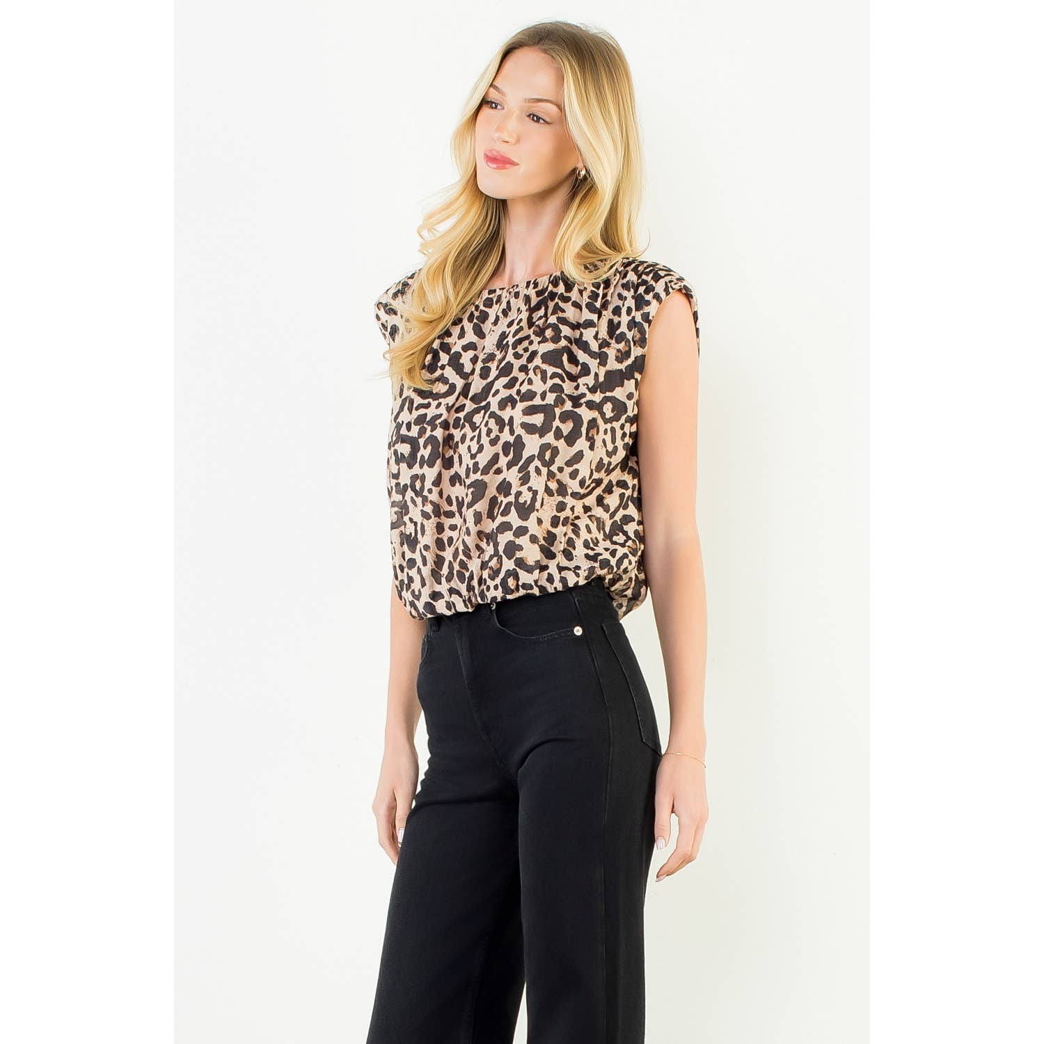 A blonde woman wearing a beige and brown leopard-print sleeveless top with black wide-leg pants, standing against a plain white studio background.