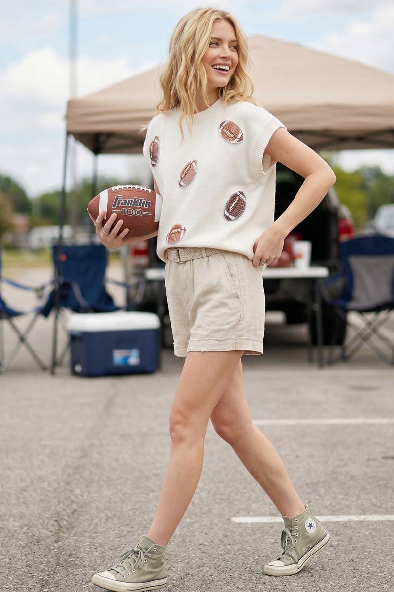A woman stands outdoors in a parking lot holding a football, wearing a cream short-sleeve knit top with football patches, beige shorts, and green sneakers. A canopy tent and folding chairs are blurred in the background.
